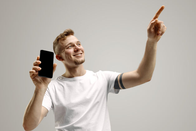 技术年轻帅哥在灰色背景下展示智能手机屏幕 一张惊喜的脸人类情感 面部表情概念惊喜商人抱