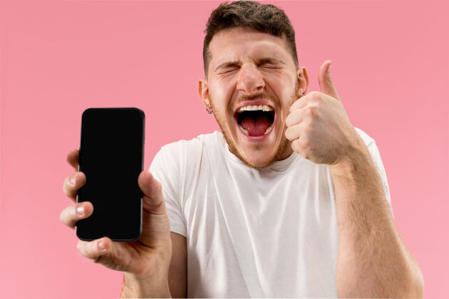 抱着年轻帅哥在粉色背景下展示智能手机屏幕 惊喜万分胡须年轻技术