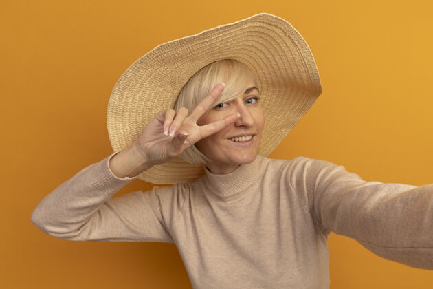 手势面带微笑的金发斯拉夫女人 戴着沙滩帽 打着胜利手势 假装拿着橙色的相机胜利成人时尚