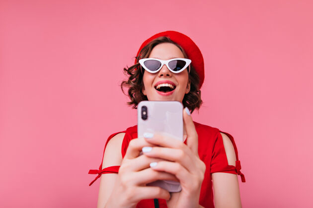 大笑一个穿着法国服装的白人女孩用手机自拍 一个戴着红色贝雷帽的黑发女士在自拍贝雷帽手机乐趣