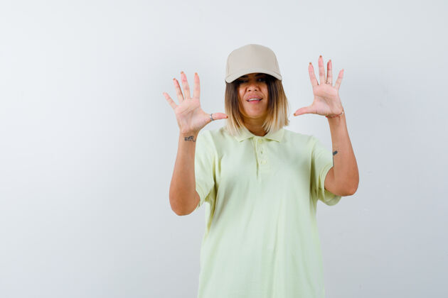 头部年轻的女士展示了在投降的手势在t恤衫 帽子和无助的样子 前视图手掌漂亮无助手掌