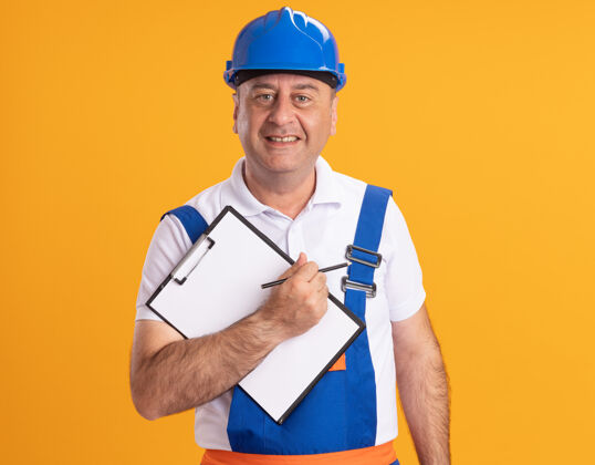 铅笔身着制服的高加索成年建设者微笑着拿着铅笔和剪贴板在橘子上建设者制服剪贴板