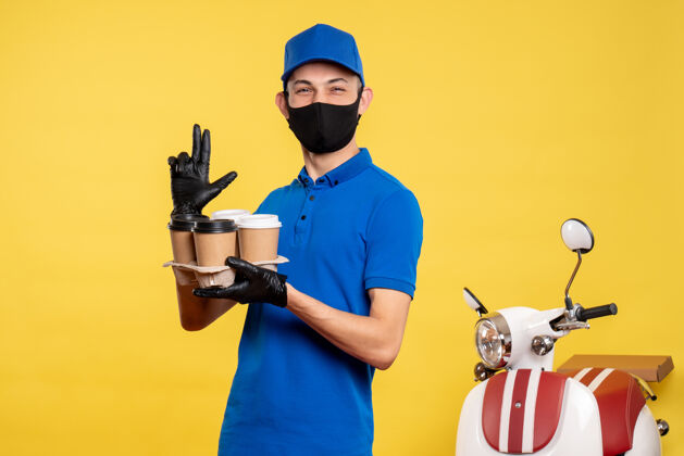 工作正面图：戴着黑色面具的男信使拿着咖啡在黄色工作服上-大流行服务递送制服工作视野持有人