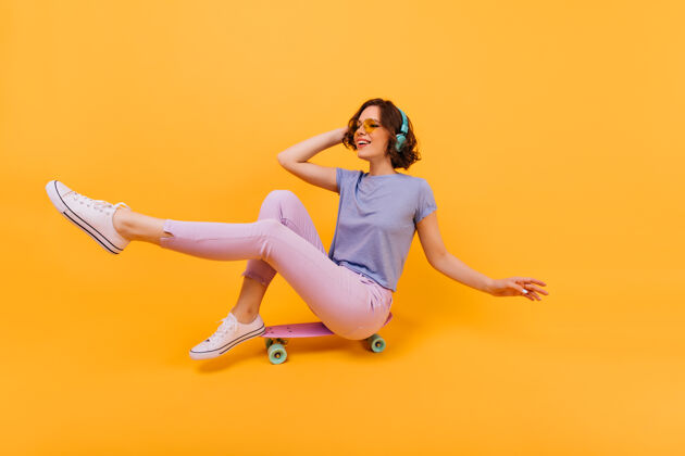 热情穿着粉色裤子的有趣女孩坐在滑板上做鬼脸戴着耳机的可爱白人女人的室内照片玩无忧无虑大笑
