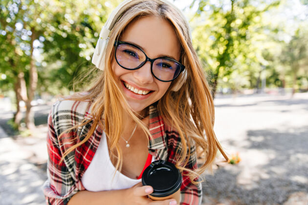 无忧无虑快乐的戴着眼镜的年轻女士在公园里喝着咖啡幸福的高加索女孩在户外享受快乐和听音乐的户外照片情感模特阳光