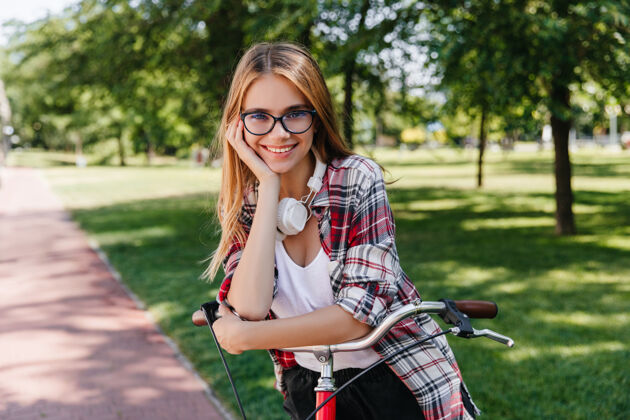 休闲热情的白人女孩戴着可爱的眼镜坐在自行车上快乐的女模特在绿色公园愉快地摆姿势的户外照片欧洲女人户外