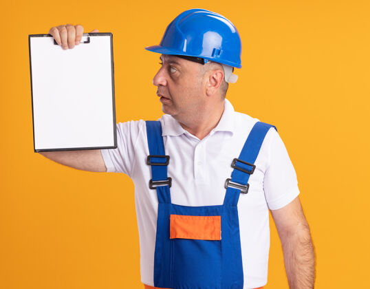 人印象深刻的白人成年建筑工人在制服举行 并在橙色的剪贴板上看印象成人人