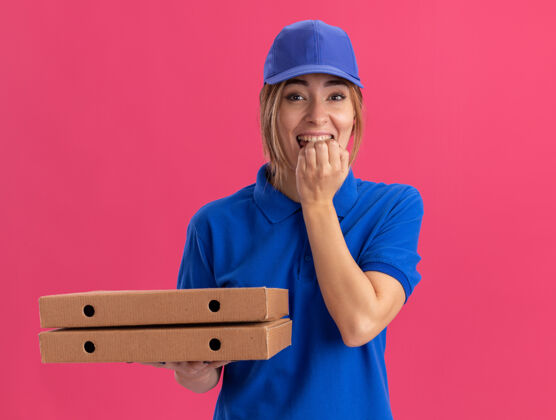 手指穿着制服的快乐的年轻漂亮的送货女孩咬着手指 拿着粉红色的比萨饼盒送货披萨制服