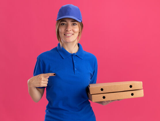 漂亮微笑着的穿着制服的年轻漂亮的送货女孩拿着并指着粉色的比萨饼盒姿势尖制服