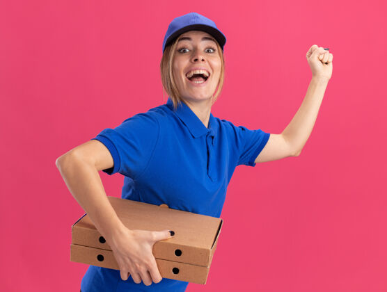 漂亮穿着制服的年轻漂亮的送货女孩兴奋地站在一边 手里拿着比萨饼盒 手里攥着粉红色的拳头侧身制服站着