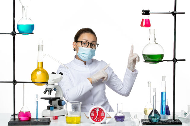 面具前视图穿着医疗服的女化学家戴着面罩坐着 溶液指向浅白色背景上的某个东西化学病毒-病毒-飞溅医生实验室解决方案