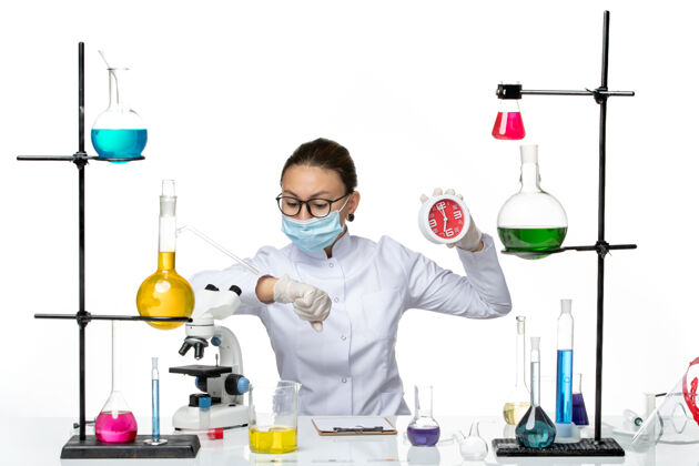 手腕前视图：穿着医疗服的女化学家戴着面具拿着时钟 看着她手腕上的白色背景病毒实验室化学冠状病毒-飞溅病毒前面医学