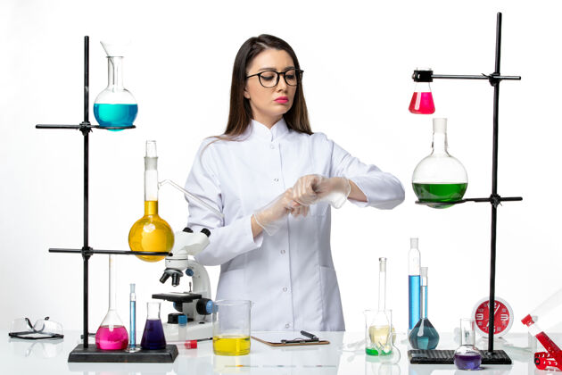 烧杯前视图：穿着无菌医疗服的女化学家在浅白背景病毒covid上看着自己的手腕-大流行科学人实验室专业