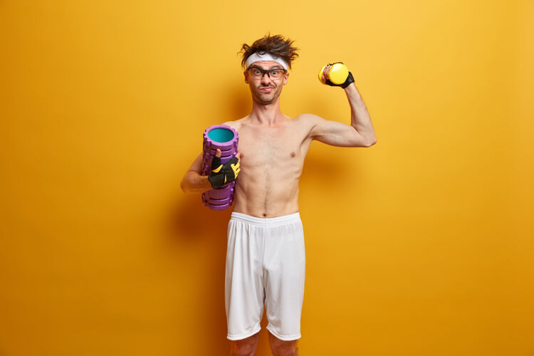 活动图为坚定的运动员举起哑铃 摆出泡沫滚轴姿势 目标是强壮 喜欢在健身房锻炼 隔离在黄色的墙壁上人 健康和健身的概念健康的生活方式健美肌肉健身房