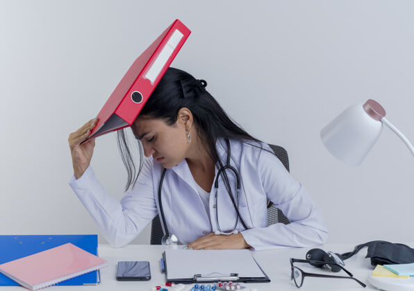 手压力大的年轻女医生穿着医用长袍和听诊器坐在办公桌旁 手持医疗工具 手放在办公桌上 用夹着头 闭着眼睛 与世隔绝桌子工具眼睛