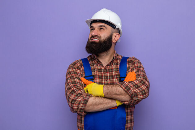 橡胶一个留着胡须的建筑工人 穿着建筑制服 戴着安全帽 戴着橡胶手套 脸上挂着微笑 脸上洋溢着幸福和自信 双臂交叉放在胸前 站在紫色的背景上施工立场制服