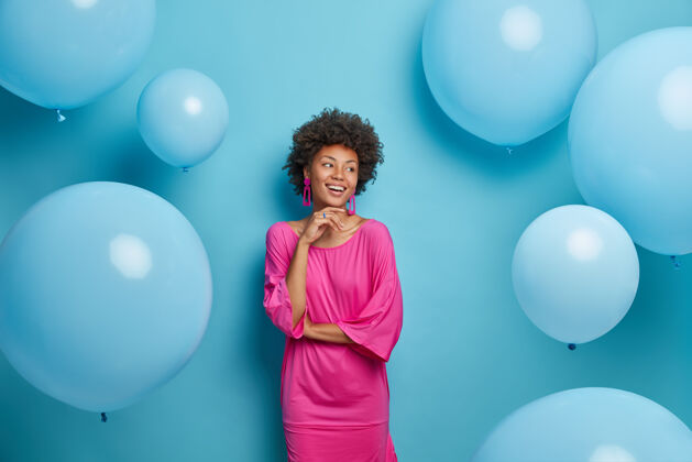 灵感快乐的年轻优雅的女士穿着粉红色的裙子享受生日聚会或其他庆祝活动 表情愉快地看着旁边 对着蓝色墙上的气球摆姿势 等待生命中的特殊事件多彩时尚心情