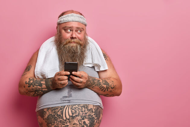 大照片中 肥胖的胡须男子在智能手机上读短信 在家里忙着健身 在体育应用程序中查看结果 他燃烧了多少卡路里 在体形过小的t恤衫上有刺青的腹部脂肪团腹部大
