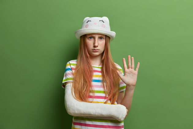 穿着严重的不高兴忧郁悲伤的女孩带着生气的表情 挥舞着手掌 向某人问好 受伤的断臂上缠着绷带 隔离在绿色的墙上孩子们受伤了姿势不高兴消极
