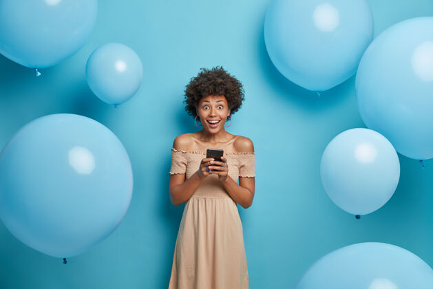 满意积极乐观的非裔美国女性手拿手机 乐于在社交网络中与朋友聊天 穿着鸡尾酒会礼服 在装饰好的照片区对着蓝色墙壁摆姿势姿势女人肖像