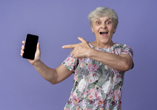 电话兴奋的老妇人拿着并指着紫色墙上的电话兴奋抱着老人