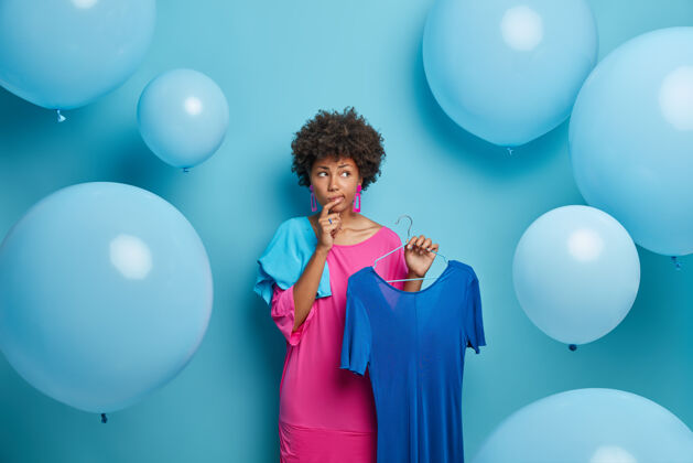 购物优柔寡断体贴的非洲裔美国妇女为宴会选择服装 考虑穿什么 把衣服挂在衣架上 用大气球隔着蓝色的墙壁女人 时尚 服装气球穿着民族