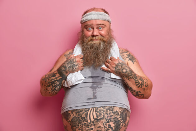 决心有趣的蓝眼睛男人的照片有大肚子 纹身的身体 忙着做健身运动 吃快餐后燃烧卡路里 隔离在粉红色的墙上大个子男人过着健康的生活胡须有氧超重