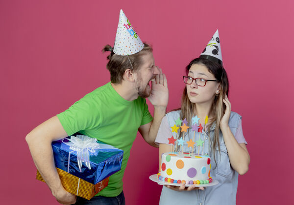 粉色恼怒的戴着派对帽的年轻人拿着礼盒 看着戴着派对帽 拿着生日蛋糕的年轻女孩大喊大叫年轻人聚会帽子