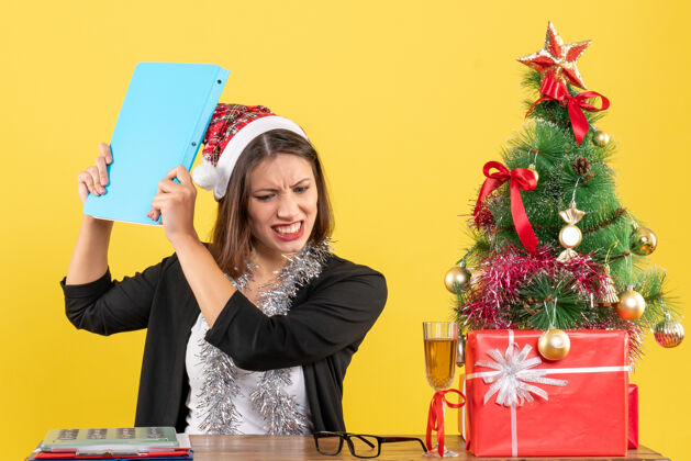 西装疲惫迷人的女士穿着西装 戴着圣诞老人的帽子和新年的装饰品 手里拿着文件 在黄色的办公室里与世隔绝快乐成人黄色