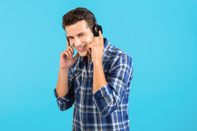 耳机时尚帅气的年轻人 戴着无线耳机听音乐 玩得开心 现代风格 快乐的情绪 蓝色背景 穿着格子衬衫无线积极享受