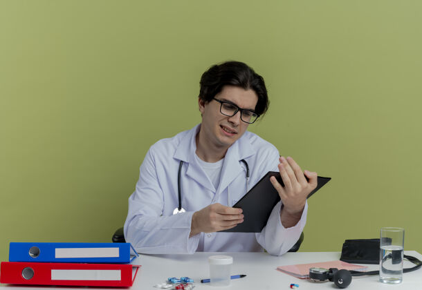 年轻人专注的年轻男医生 穿着医用长袍 戴着听诊器 戴着眼镜 坐在办公桌旁 拿着医疗工具 孤立地看着剪贴板工具穿着桌子