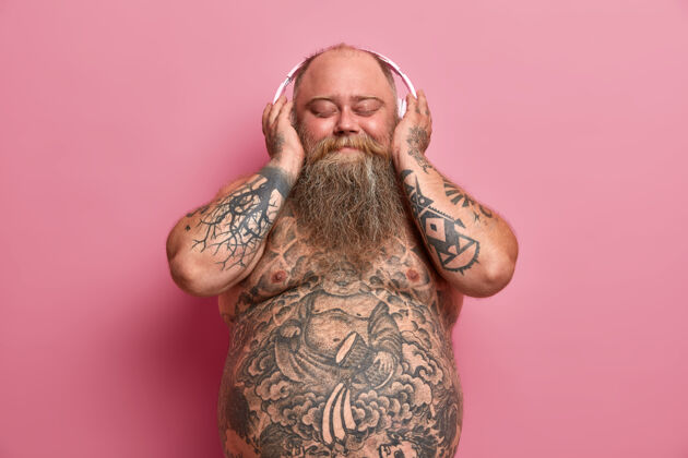 音乐快乐肥胖的胖子喜欢用立体声耳机听喜爱的音乐 裸着肚子摆姿势 手臂和肚子上有纹身 因为吃快餐而超重 隔离在粉红色的墙上身体表情丰满