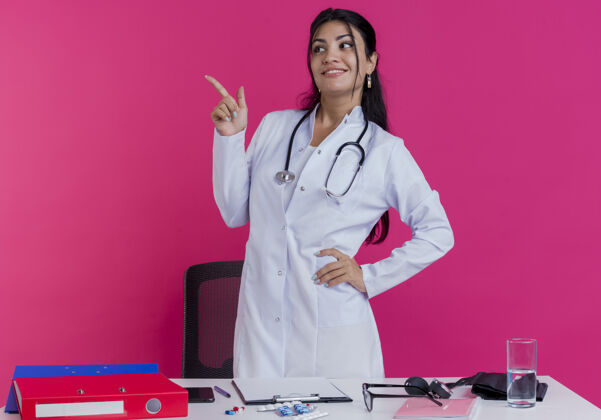 保持面带微笑的年轻女医生穿着医用长袍 手持听诊器站在桌子后面 手持医疗工具 手放在腰上 看着并指着隔离的一侧女医生年轻人