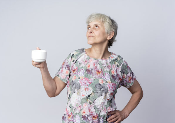 拿着高兴的老妇人拿着杯子 把手放在腰上 孤零零地看着白墙上起来杯子老的