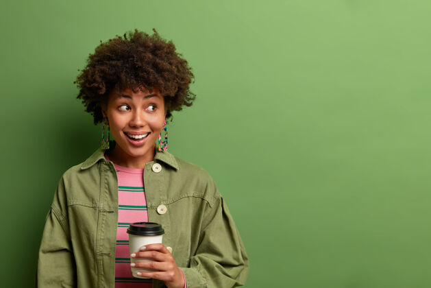 非洲快乐的黑皮肤女人拿着一次性的热饮杯 一边看一边高兴地笑着 喝咖啡休息 享受早晨的饮料 隔离在绿色的墙上 复制空间刷新年轻个人