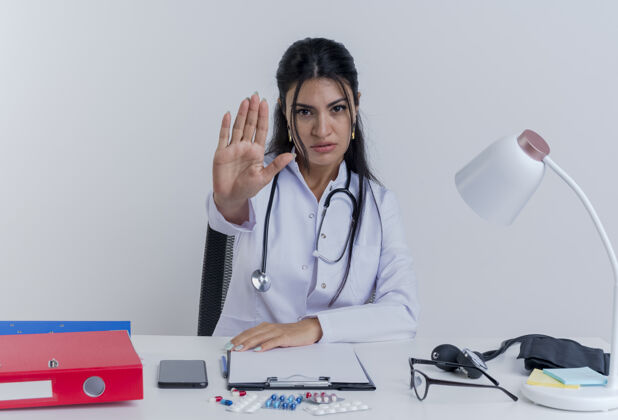 人严格的年轻女医生穿着医用长袍和听诊器坐在办公桌旁用医疗工具把手放在办公桌上看着做停止手势医生坐着医疗