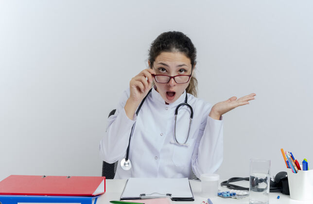 桌子令人印象深刻的年轻女医生穿着医用长袍和听诊器 戴着眼镜坐在办公桌旁 拿着医疗工具 抓起眼镜 空手看起来很孤立长袍医疗空的