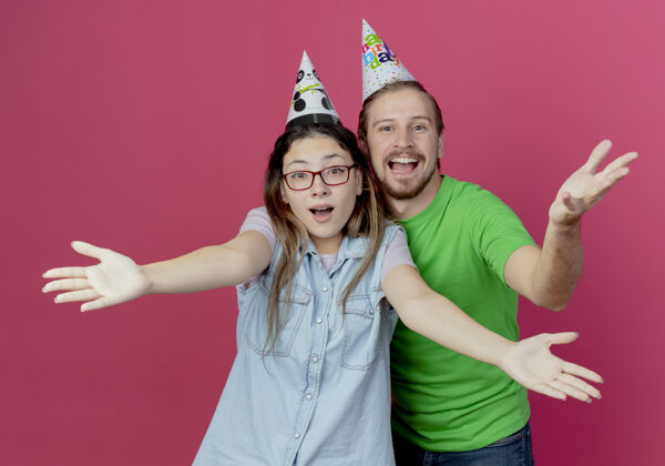 手快乐的年轻夫妇戴着派对帽 在粉红色的墙上举手致意情侣快乐穿