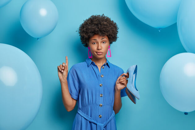 优雅严肃自信的女人点以上邀请你上楼拿着新鞋穿高跟鞋穿着时髦的衣服 试穿衣服出门 摆着气球对着蓝色的墙壁女人指向尝试