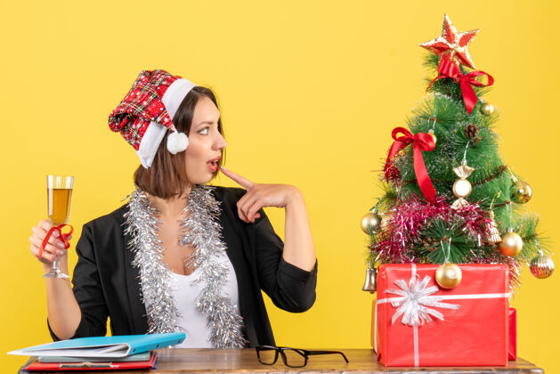 肖像想着迷人的女士 穿着西装 戴着圣诞老人的帽子和新年的装饰品 手里拿着红酒 在黄色的办公室里与世隔绝帽子圣诞老人人