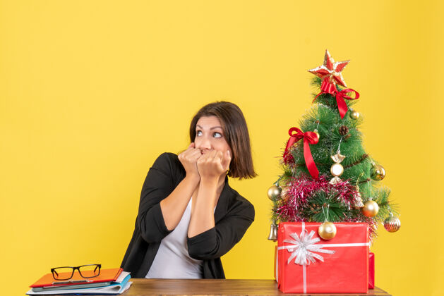 人在黄色的办公室里 一个害怕的年轻女人坐在装饰过的圣诞树旁的桌子旁看着什么东西肖像树女士