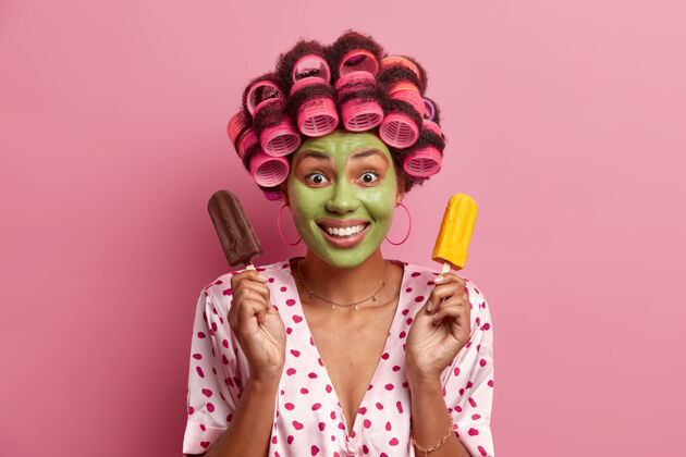 冰淇淋好看的女模特肖像在脸上涂上绿色的美容面膜 戴上卷发器做卷发 拿着巧克力和芒果冰淇淋 心情愉快 笑容灿烂 孤立在粉色上高兴家庭主妇发型