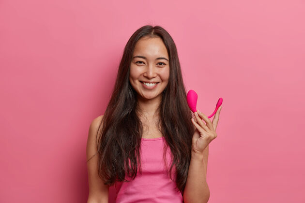 情绪快乐的黑发女人用智能蓝牙振动器摆出姿势 用手机上的特殊应用程序来提高性高潮 拿着性爱工具来增强快感 隔离在粉红色的墙上现代科技和性生活高兴性现代