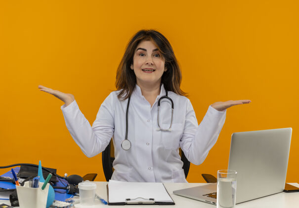 人令人印象深刻的中年女医生穿着医用长袍和听诊器坐在办公桌前 医疗工具剪贴板和笔记本电脑显示空手孤立长袍女士成人
