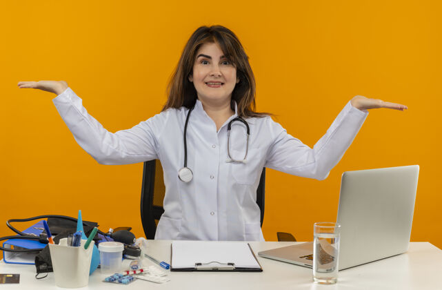 穿快乐的中年女医生穿着医用长袍和听诊器坐在办公桌前 医疗工具剪贴板和笔记本电脑显示空手孤立医疗桌子医生