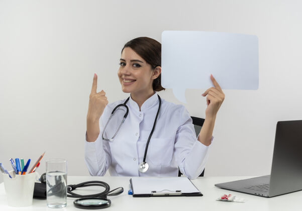 女士面带微笑的年轻女医生穿着医用长袍和听诊器坐在办公桌旁 手里拿着医疗工具和笔记本电脑 手里拿着聊天泡泡 指指点点女性起来听诊器