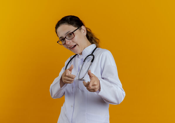 表演高兴的年轻女医生穿着医用长袍 戴着眼镜听诊器给你看听诊器成人女士
