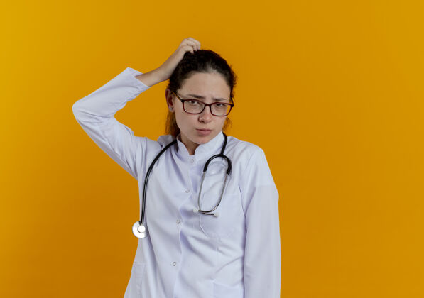刮迷茫的年轻女医生穿着医用长袍 戴着眼镜听诊器搔头孤立医生眼镜年轻