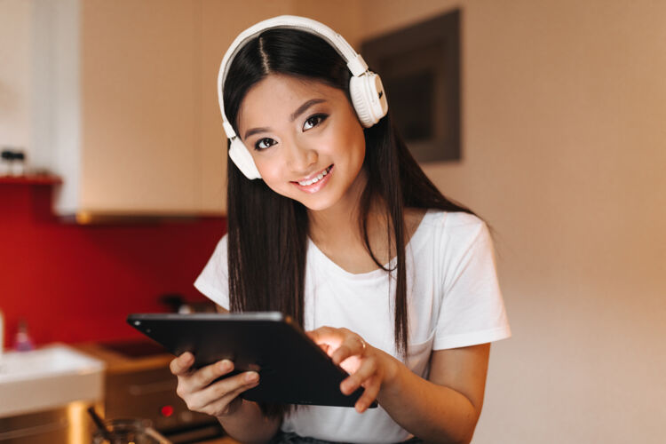 设备棕色眼睛的女人戴着宽大的耳机看着前面 微笑着拿着平板电脑公寓房子互联网