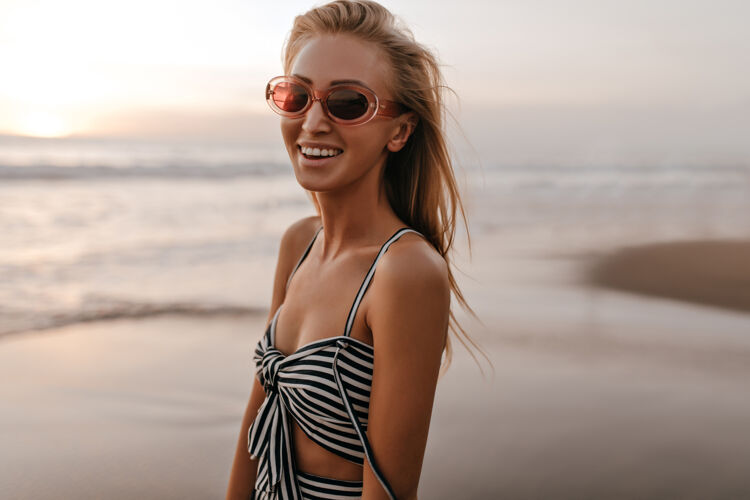 春天美丽快乐的金发女郎 戴着时髦的粉色太阳镜 穿着不规则的条纹上衣 在日落时分微笑着走在沙滩上时尚发髻人
